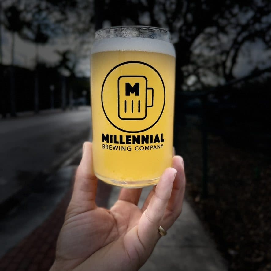 A hand holding a Millennial Brewing Company beer glass on a downtown Fort Myers street at dusk